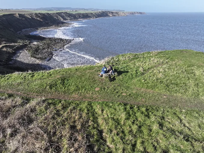 Two walkers resting on the clifftop path overlooking the Yorkshire Coast near Scarborough in autumn, with dramatic cliffs, rocky shoreline and the vast sea below